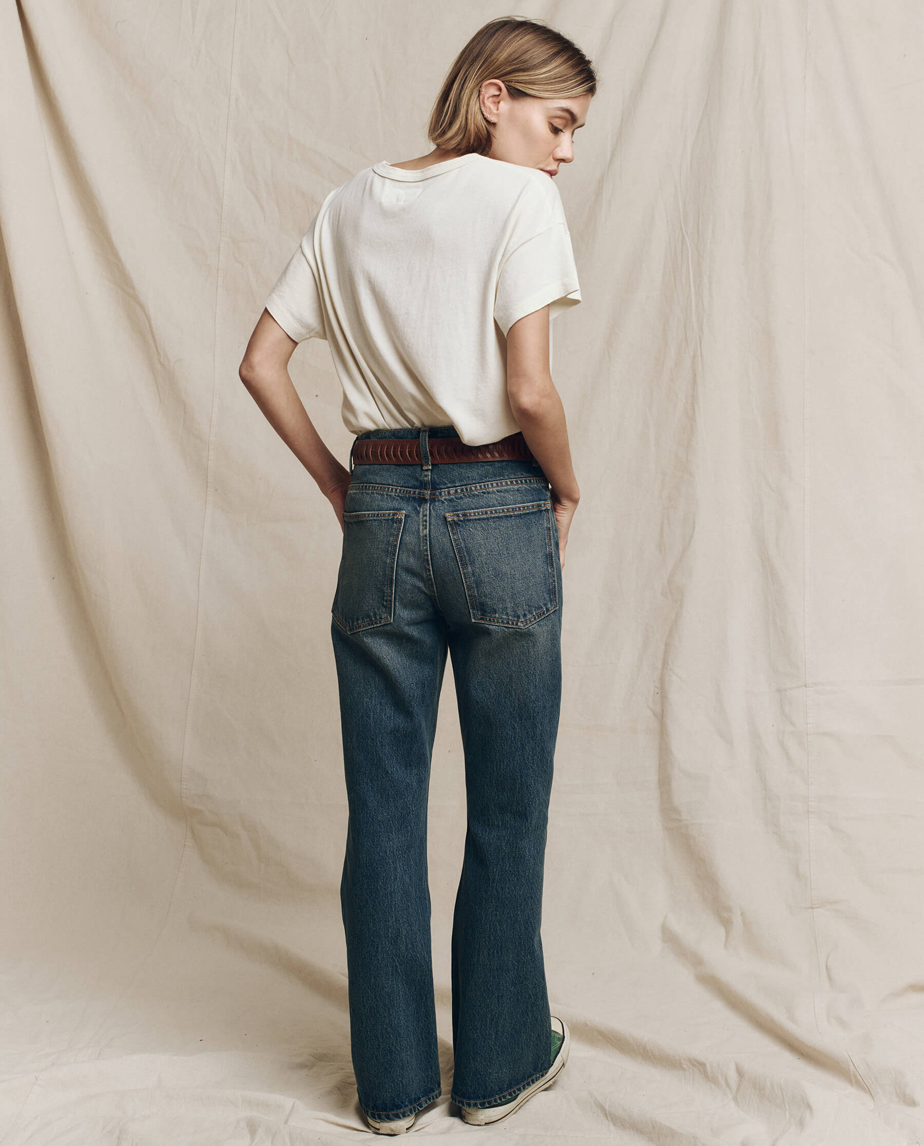 A person stands with their back turned, wearing a Washed White Limited Edition: The I Am A Voter Boxy Crew T-shirt by The Great. and blue jeans, set against a plain, beige fabric backdrop.