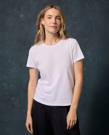 A woman with long blonde hair wears The Little Tee. Solid -- True White by THE GREAT. SP22 APRILCAP and dark pants, standing before a dark mottled background.
