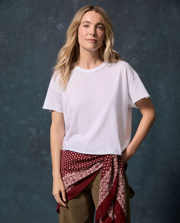 A person with long blonde hair wears The Crop Tee. Solid -- True White by THE GREAT. CORE KNITS, olive pants, and a red patterned scarf at the waist against a dark blue background.