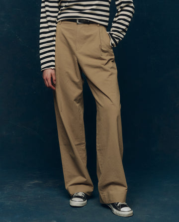 Wearing THE GREAT. PS26 COLLECTORS The Menswear Trouser in khaki, a black-and-white striped long-sleeve shirt, and matching sneakers, the person stands against a dark background with their face not visible.