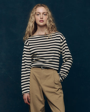 A person with long, wavy blonde hair wears THE GREAT. SP25 COLLECTORS The Sailor Sweater -- Black Stripe with beige pants, standing against a dark blue background.
