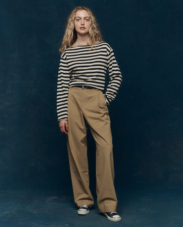 A person stands facing forward against a dark blue background, wearing THE GREAT. SP25 COLLECTORS The Sailor Sweater in Black Stripe, tan wide-leg pants, and black sneakers.