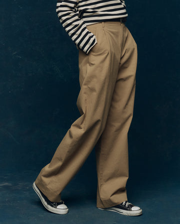 A person stands against a dark background wearing The Menswear Trouser in khaki by THE GREAT. PS26 COLLECTORS, paired with a black-and-white striped long-sleeve shirt and black low-top sneakers.
