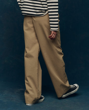 A person wearing a black-and-white striped long-sleeve shirt, THE GREAT. PS26 COLLECTORS The Menswear Trouser in Khaki, and black sneakers stands against a dark background.