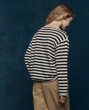 A person with long hair, wearing THE GREAT. SP25 COLLECTORS The Sailor Sweater in Black Stripe and beige pants, stands facing away against a dark blue background.