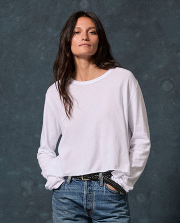 A woman with long brown hair wears The Long Sleeve Crop Tee. -- True White by THE GREAT. CORE KNITS and blue jeans, standing against a dark background with hands in her pockets.