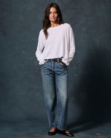 A person stands with hands in pockets, wearing THE GREAT. CORE KNITS The Long Sleeve Crop Tee. -- True White, blue raw-hem jeans, black belt, and black flats against a dark background.