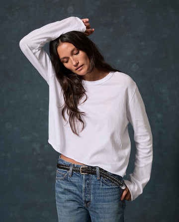 Wearing THE GREAT. CORE KNITS The Long Sleeve Crop Tee. -- True White and blue jeans, a person stands against a dark background, arm bent and head slightly lowered.