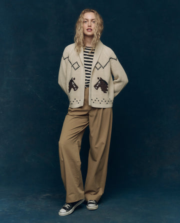 Person standing against a dark backdrop, wearing a cream cardigan with animal designs, a black and white striped shirt, THE GREAT. PS26 COLLECTORS The Menswear Trouser in khaki, and black sneakers.