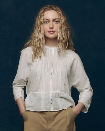 A person with long curly blond hair wears THE GREAT. F25 COLLECTORS The Victorian Top in white and tan pants, standing against a dark blue background.