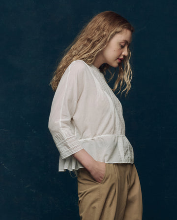 A woman with long, wavy blonde hair stands in profile against a dark blue background, wearing THE GREAT. F25 COLLECTORS' The Victorian Top in white and tan pants.