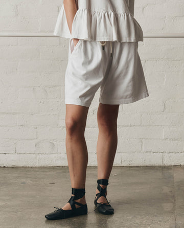 Indoors, a person wears THE GREAT. SP26 DANCER BODIES’ The Relaxed Sport Short in True White—featuring an elastic waistband and pockets—with a white ruffled top and black lace-up flats. The backdrop is a light brick wall and concrete floor.