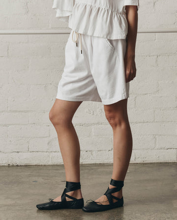 A person stands indoors against a white brick wall wearing THE GREAT.'s True White Relaxed Sport Short and a white ruffled top, paired with black lace-up flats.