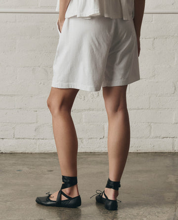 A person stands indoors wearing THE GREAT.'s The Relaxed Sport Short in True White, paired with a white top and black ballet flats with ribbon ankle ties. Only the lower half of the body is shown.