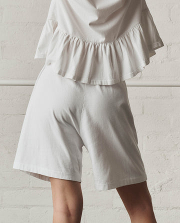 A person wearing THE GREAT. SP26 DANCER BODIES The Relaxed Sport Short in True White, featuring an elastic waistband, and a white ruffled top stands against a light-colored brick wall with their upper body partially out of frame.