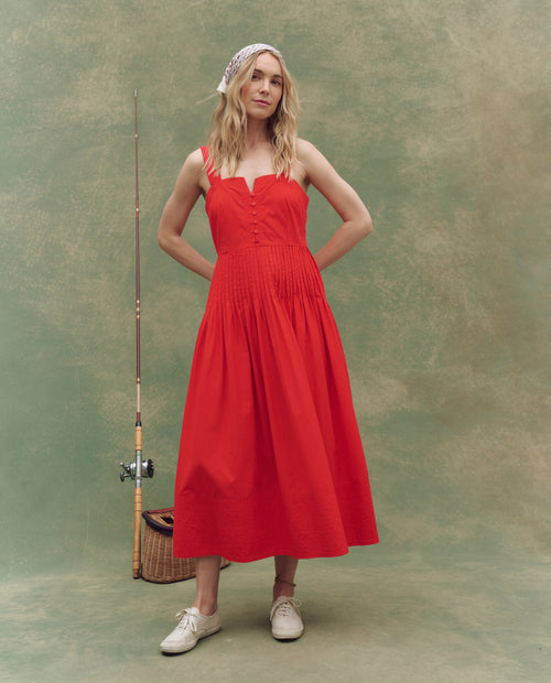 A woman in THE GREAT. SU25's Hot Red The Day Break Dress and white shoes stands indoors on a green backdrop, holding a fishing rod and wearing a patterned headscarf—a perfect choice for casual women’s fashion.