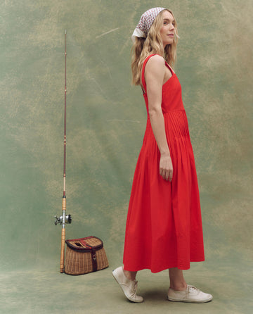 A woman stands sideways in THE GREAT. SU25 The Day Break Dress in Hot Red with white sneakers and a headscarf, beside a fishing pole and wicker bag against a neutral green backdrop—an effortless fashion statement.