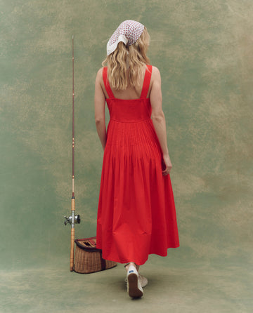 Wearing THE GREAT. SU25 The Day Break Dress in Hot Red and a white headscarf, a person stands with their back to the viewer beside a fishing rod and wicker basket, set against a mottled green backdrop—evoking laid-back style.