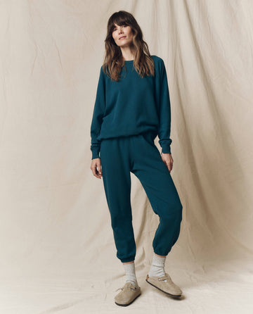 In Los Angeles, a person stands before a beige fabric backdrop wearing THE GREAT. F25 SEASONAL KNITS The Stadium Sweatpant. Solid -- Dark Slate, styled with a teal sweatshirt, gray socks, and tan slip-on shoes.