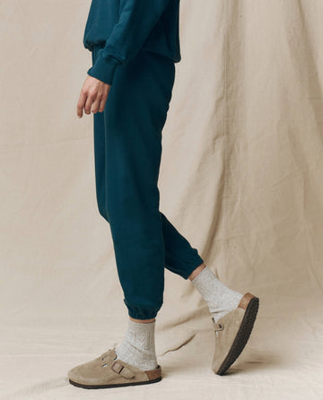 Person wears The Stadium Sweatpant. Solid -- Dark Slate by THE GREAT. F25 SEASONAL KNITS, paired with beige socks and slip-on sandals, standing before a beige fabric backdrop for a relaxed Los Angeles vibe.