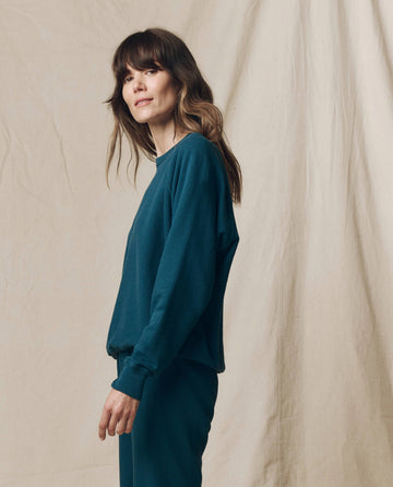 Woman standing sideways, wearing THE GREAT. F25 SEASONAL KNITS "The College Sweatshirt. Solid -- Dark Slate," styled as a matching skirt set, with a neutral beige backdrop—a handcrafted Los Angeles vibe.