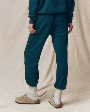 A person stands on a light fabric backdrop wearing THE GREAT. F25 SEASONAL KNITS The Stadium Sweatpant in Dark Slate with a matching top, beige socks, and light brown slip-on sandals—a relaxed Los Angeles-inspired look.