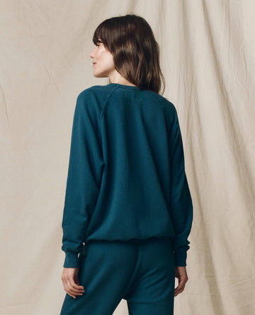 A person with brown hair wears THE GREAT. F25 SEASONAL KNITS The College Sweatshirt, Solid in Dark Slate, facing away from the camera against a beige fabric backdrop.