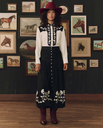 Wearing THE GREAT. F26 STETSON’s The Color Block Embroidered Western Shirt in cream with black, a person in a western-style outfit and red hat stands indoors before a wall of framed horse paintings on a dark background, their crisp shirt adding to the authentic cowboy flair.