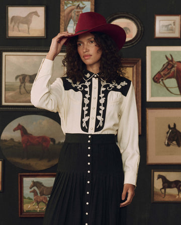 Wearing THE GREAT. F26 STETSON’s The Color Block Embroidered Western Shirt in cream with black, a woman in a striking red hat stands before a wall adorned with framed paintings of horses.