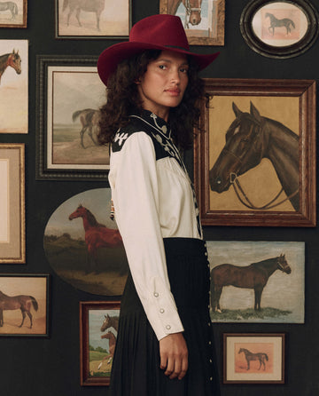 Modeling THE GREAT. F26 STETSON’s The Color Block Embroidered Western Shirt in Cream with Black, a person pairs it with a black skirt and a red cowboy hat while standing before a wall adorned with framed horse paintings and photographs.