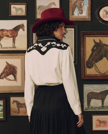 Wearing The Color Block Embroidered Western Shirt in cream with black by THE GREAT. F26 STETSON, a person in a red hat stands with their back to the camera, surrounded by framed horse paintings on a dark wall—creating a striking tableau that captures the spirit of this unique collaboration.