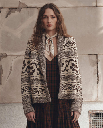 Wearing THE GREAT. F26 D1’s The Nordic Flower Lodge Cardigan in Marled Chestnut, a woman with wavy hair stands against a weathered stone wall. She pairs the patterned brown-and-white cardigan with a dark plaid dress and a light-colored neckerchief.