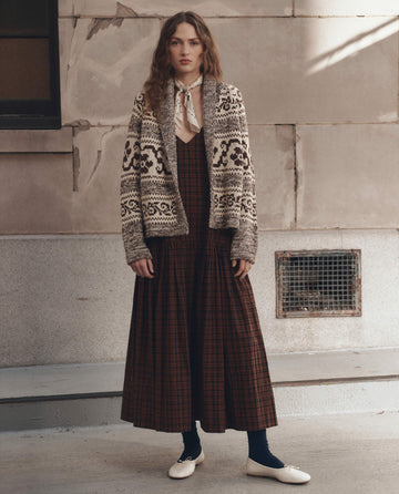 A woman stands outside in front of a beige wall, wearing THE GREAT.'s The Nordic Flower Lodge Cardigan in Marled Chestnut over a long plaid dress, paired with navy tights, white flats, and a light scarf tied around her neck.