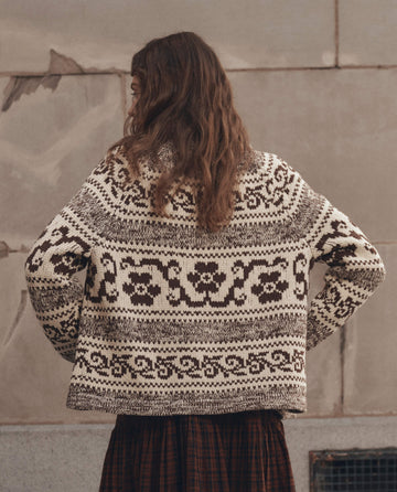 A person with long, wavy brown hair seen from behind is wearing THE GREAT. F26 D1's The Nordic Flower Lodge Cardigan in Marled Chestnut, featuring floral and geometric patterns in brown and cream, while standing in front of a textured stone wall.