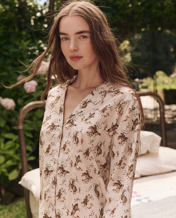 A woman with long brown hair stands outdoors amid greenery and flowers, wearing THE GREAT. SP25 SLEEP's The Pajama Shirt in Cream Cowboy, a light-colored, animal print sleepwear piece.