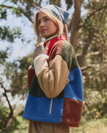 A woman in THE GREAT. SP26 TGO Plush Color Block Sierra Pullover in Canyon Multi and a blue headscarf stands outdoors with trees behind her.