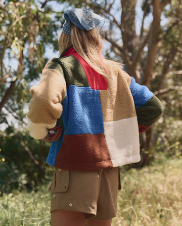 A person stands outdoors facing away, wearing THE GREAT. SP26 TGO The Plush Color Block Sierra Pullover in Canyon Multi, khaki floral-detailed shorts, and a blue patterned headscarf.