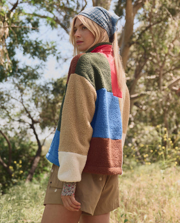Wearing THE GREAT. SP26 TGO The Plush Color Block Sierra Pullover in Canyon Multi, khaki shorts, and a blue headscarf, a person stands outdoors among trees and grass, looking over their shoulder.