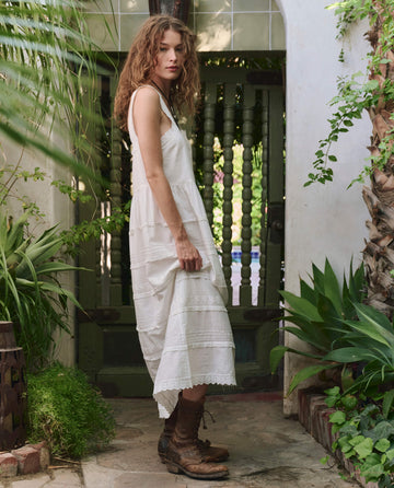 A woman in THE GREAT. PF25 The Reverie Dress—White, paired with brown boots, stands on a stone path by a green gate amid lush plants, embodying effortless style in a tranquil setting.