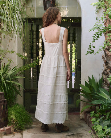 A woman with wavy hair, dressed in THE GREAT. PF25's The Reverie Dress in white and brown shoes, stands outdoors facing a green wooden gate amidst lush plants.