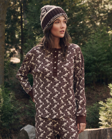 A woman in brown and cream pajamas and a knit hat stands outdoors by green trees beside The Slope Henley. -- Umber from THE GREAT. F26 TGO, looking to the side.