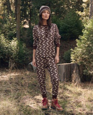 A person stands outdoors among trees and grass, wearing The Slope Henley. -- Umber by THE GREAT. F26 TGO with a brown patterned outfit, beanie, and red hiking boots.