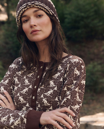 A woman in The Slope Henley. -- Umber by THE GREAT. F26 TGO stands outdoors with arms crossed, exuding relaxed, casual style against a backdrop of blurred greenery.
