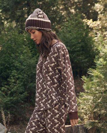 Wearing The Slope Henley. -- Umber by THE GREAT. F26 TGO, a person strolls outdoors in patterned brown knit accessories and bear-print pajamas near green trees.