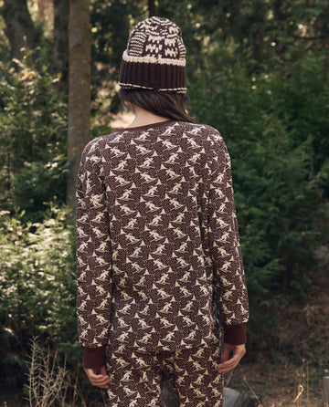 Person standing outdoors, back to camera, wearing The Slope Henley. -- Umber by THE GREAT. F26 TGO under a brown patterned sweater, with matching pants and a knit hat. Trees in background.