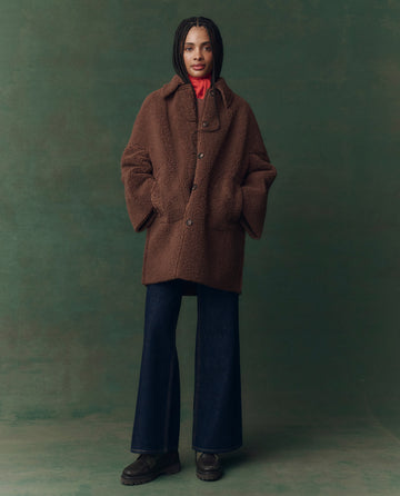 A person stands facing forward in THE GREAT. F25 D2 The Plush Car Coat in chocolate, paired with a red hoodie, wide-leg dark jeans, and black shoes against a green background.