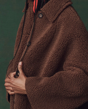 A person wears THE GREAT. F25 D2's The Plush Car Coat in chocolate, styled with a 1950s silhouette and large buttons, holding the front closed with one hand against a solid green background.