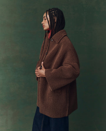A person with braided hair wears THE GREAT. F25 D2’s The Plush Car Coat in chocolate, styled with dark pants and shown in profile against a solid green background.