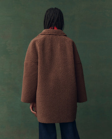 A person with braided hair, wearing THE GREAT. F25 D2's The Plush Car Coat in Chocolate and dark pants, stands facing away from the camera against a green background.