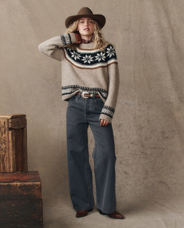 A person wearing THE GREAT. H25 COLLECTION's The Snowflake Fairisle Pullover in Oat stands indoors near wooden crates against a neutral backdrop, styled with a brown hat, wide-leg jeans, and brown boots.